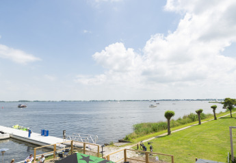 Vista desde una casa flotante hacia un lago con barcos, muelle, césped verde y cielo parcialmente nublado.