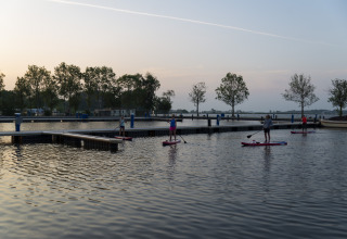 Menschen beim Stand-Up-Paddling nahe dem Hausboot Aqua Dolce in Friesland, Niederlande, bei Sonnenuntergang.