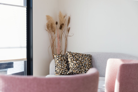 Rincón luminoso de salón en una casa flotante con sofá gris, cojines geométricos y sillas rosas.