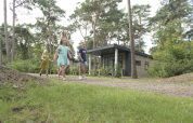 Familia paseando frente al Forest park chalet en Holiday park De Kleine Belties, rodeada de árboles en los Países Bajos.