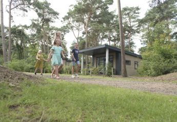 Familia paseando frente al Forest park chalet en Holiday park De Kleine Belties, rodeada de árboles en los Países Bajos.