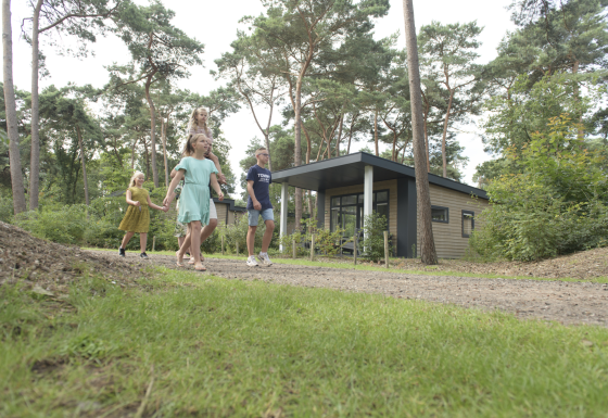 Famille marchant devant le Forest park chalet à Holiday park De Kleine Belties, entouré d’arbres aux Pays-Bas.