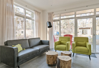 Modern living room with large windows, a grey sofa, two green armchairs, and log tables at Forest park chalet.