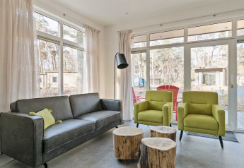 Moderne woonkamer met grote ramen, grijze bank, twee groene stoelen en boomstamtafels in Forest park chalet.