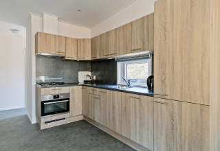 Modern kitchen interior with wood finish, oven, gas stove, and window at Forest park chalet, De Kleine Belties.