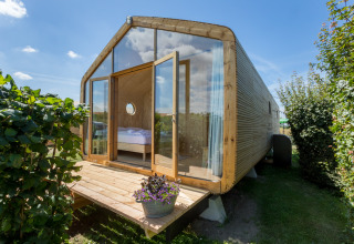 Wikkelhouse con fachada acristalada y porche de madera, rodeado de vegetación en Welcome In - Zeeland.