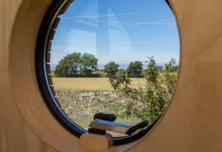 Vista a través de una ventana redonda en una Wikkelhouse en Welcome In - Zeeland, Países Bajos, con campos.