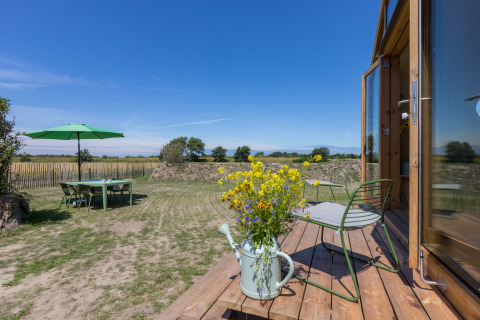Vista desde un Wikkelhouse en Zeeland, Países Bajos, con terraza, silla verde, flores y mesa de jardín.