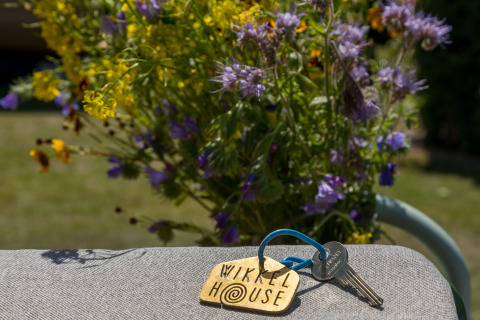 Llaves con etiqueta 'Wikkelhouse' sobre una silla, flores silvestres detrás, Welcome In - Zeeland, Países Bajos.
