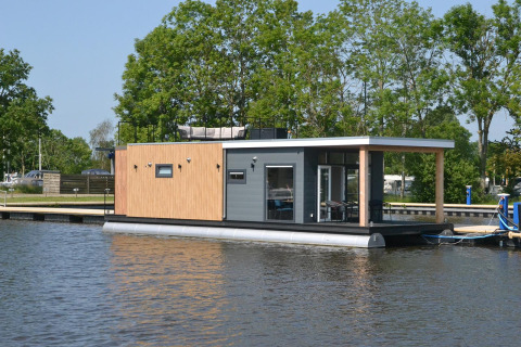 Casa flotante 'Riggelbrug' en Welcome In - Friesland, Países Bajos, atracada junto a un embarcadero y árboles.