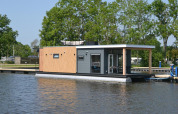 Moderne husbåd 'Riggelbrug' fortøjet ved Welcome In - Friesland omgivet af vand og træer i Holland.