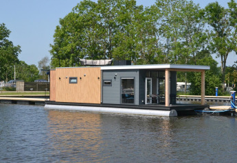 Modern houseboat 'Riggelbrug' at Welcome In - Friesland, Netherlands, floating near a dock with trees behind.