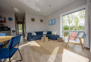 Moderne woonkamer in Houseboat 'Riggelbrug' met groot raam, blauwe zetel en uitzicht op het kanaal in Friesland.