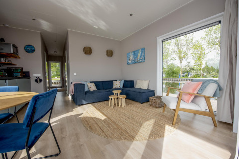 Salón moderno en la casa flotante 'Riggelbrug' con sofá azul, gran ventana y vistas al canal en Friesland.