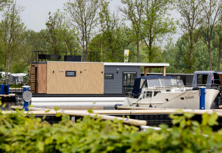 Woonboot 'Riggelbrug' bij Welcome In - Friesland, Nederland, aangemeerd naast een boot in een groene jachthaven.