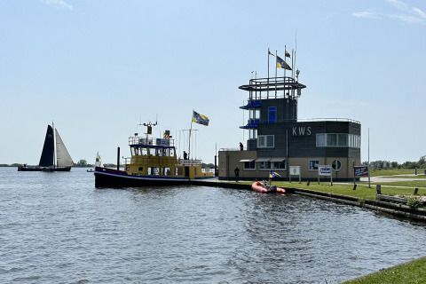 Casa flotante y edificio de control junto al agua en Friesland, Países Bajos, con banderas y veleros.
