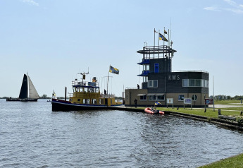 Casa flotante y edificio de control junto al agua en Friesland, Países Bajos, con banderas y veleros.