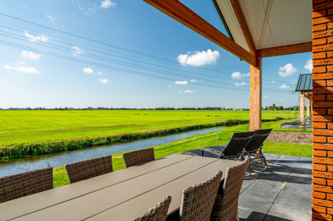 Vue du lodge Anker à Tusken de Marren aux Pays-Bas avec terrasse, champs verts, canal et ciel ensoleillé.