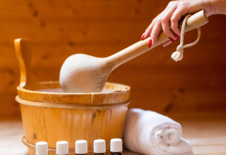 Una mano agita una cuchara de madera en una sauna, con aceites esenciales y una toalla enrollada delante.