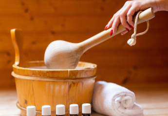 Una mano mescola un mestolo di legno in sauna, con oli essenziali e un asciugamano arrotolato davanti.