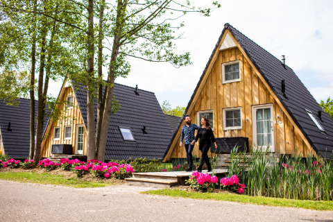 Una coppia cammina davanti alla tiny house Wellness Lodge presso Hof van Salland, Paesi Bassi, tra fiori e alberi.