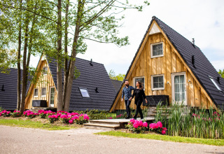 Una coppia cammina davanti alla tiny house Wellness Lodge presso Hof van Salland, Paesi Bassi, tra fiori e alberi.