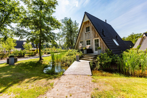 Tiny Wellness Lodge au Hof van Salland, Pays-Bas, avec façade bois, étang et passerelle bordée de verdure.