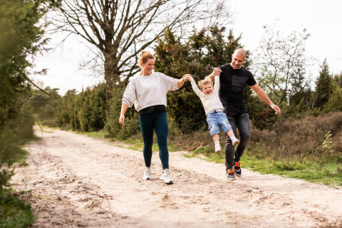 Genitori sorridenti fanno oscillare il figlio mentre camminano su un sentiero sabbioso vicino a Wellness Lodge, Hof van Salland, Paesi Bassi.