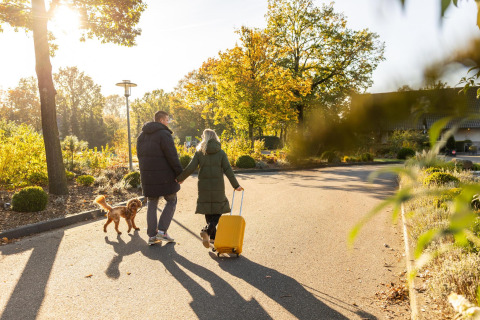 Par går hånd i hånd med hund og gul kuffert ved Wellness Lodge, Hof van Salland, Nederlandene i solskin.