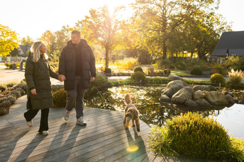 Pareja paseando a su perro junto a un estanque en Wellness Lodge, Hof van Salland, Países Bajos, soleado.