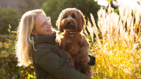 Kvinde med blondt hår holder brun hund foran Wellness Lodge ved Hof van Salland, omgivet af sollys.