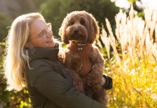 Mujer rubia sostiene a un perro marrón frente al Wellness Lodge en Hof van Salland, iluminados por el sol.