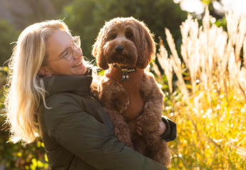 Mujer rubia sostiene a un perro marrón frente al Wellness Lodge en Hof van Salland, iluminados por el sol.