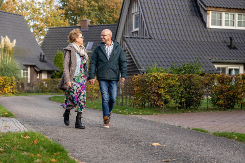 Et ældre par går hånd i hånd på en sti foran Wellness Lodge ved Hof van Salland, Holland, omgivet af hytter.