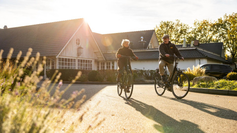 To ældre voksne cykler foran Wellness Lodge ved Hof van Salland i Holland, solskin og have omgiver dem.