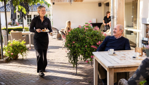 En tjener serverer dessert til en ældre mand, der nyder kaffe udendørs ved Wellness Lodge, Hof van Salland.
