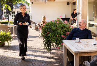 Una camarera lleva postre a un hombre mayor tomando café al aire libre en Wellness Lodge, Hof van Salland.