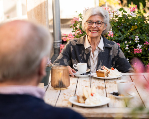 Pareja mayor disfruta café y pastel al aire libre en Wellness Lodge, Hof van Salland, rodeados de flores.