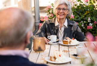 Coppia anziana gusta caffè e torta all'aperto al Wellness Lodge, Hof van Salland, tra fiori colorati.