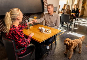 Un couple savoure boissons et dessert à table, avec un chien brun à côté au Wellness Lodge, Hof van Salland.