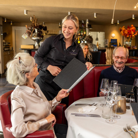 En tjener smilende serverer et ældre par i en hyggelig restaurant med rød polstrede stole i Holland.