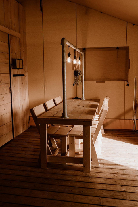 Mesa de madera con bancos y bombillas colgantes en una tienda safari con iluminación cálida e interior de madera.