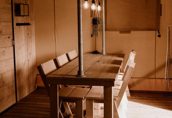 Holztisch mit Bänken und Hängelampen in einem Safari-Zelt mit warmer Lichtstimmung und Holzboden.