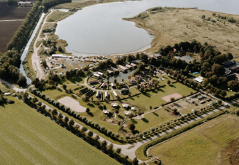 Luftfoto af et glampingområde med telte, sø, grønne marker og rekreative faciliteter i naturen.