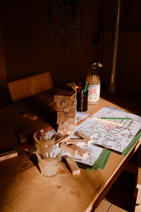 Jenga-Steine, Malstifte, Saftglas und Ausmalbilder auf einem Holztisch in einem Safari-Zelt bei warmem Licht.