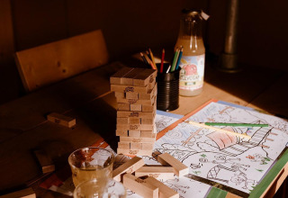 Jenga-Steine, Malstifte, Saftglas und Ausmalbilder auf einem Holztisch in einem Safari-Zelt bei warmem Licht.