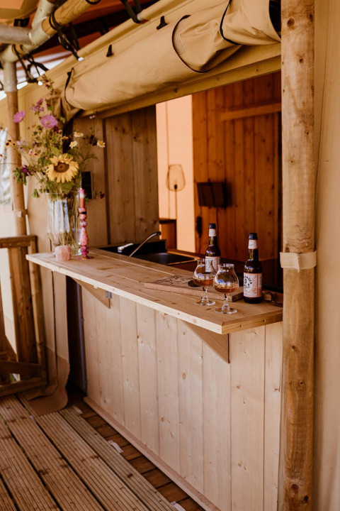 Bar extérieur dans une tente safari, avec comptoir en bois, fleurs, bouteilles de bière et verres visibles.