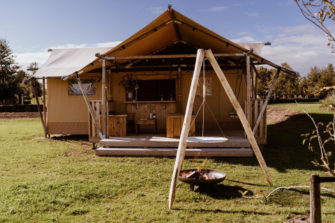 Tente safari avec terrasse en bois, balançoire et brasero, sur pelouse verte sous ciel bleu.
