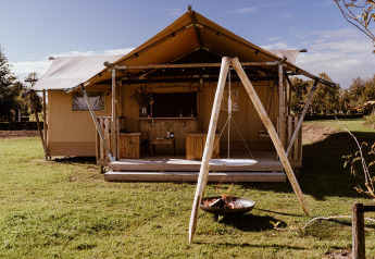 Safari-Zelt mit Holzterrasse, Schaukel und Feuerstelle auf grünem Rasen unter blauem Himmel.