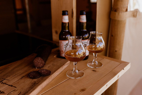 Deux verres et bouteilles de bière avec saucisson sur table en bois dans une tente safari, ambiance détendue.
